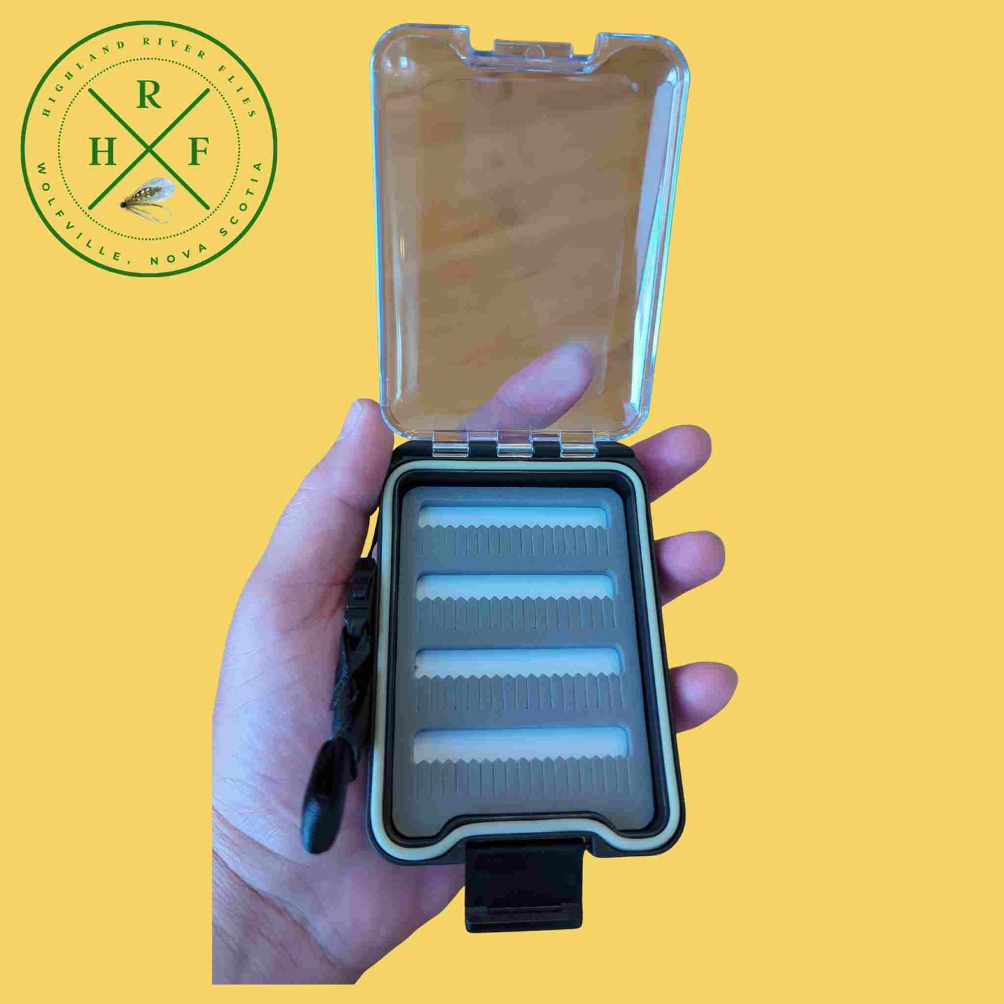 Hand holding a fishing fly box with a yellow background and a logo in the corner.